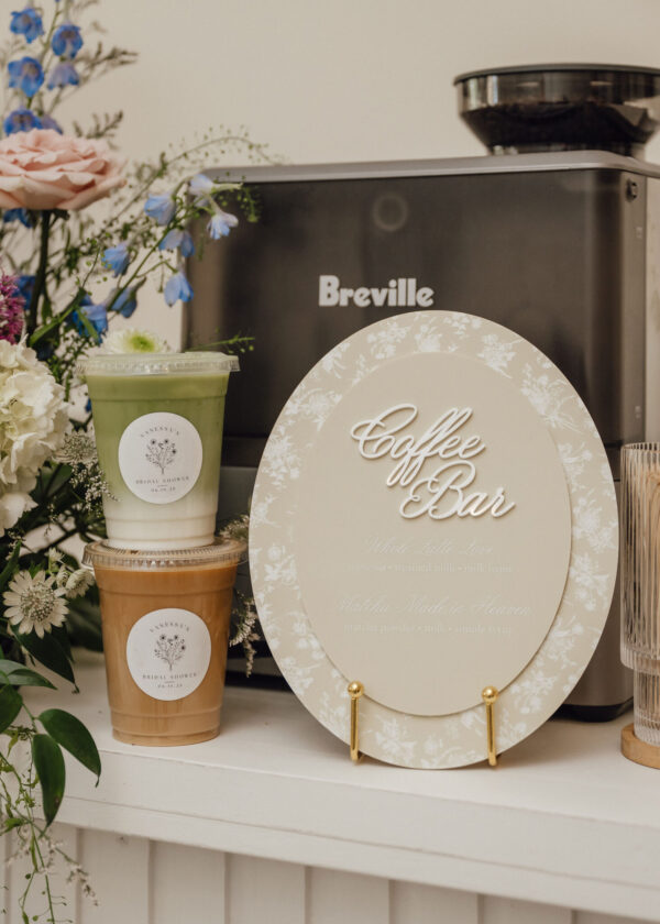 Display scene of a Coffee Bar setup with a Breville machine, decorative flowers, and two takeaway cups (green tea/juice) in front of a decorative circular sign reading 'Coffee Bar'.