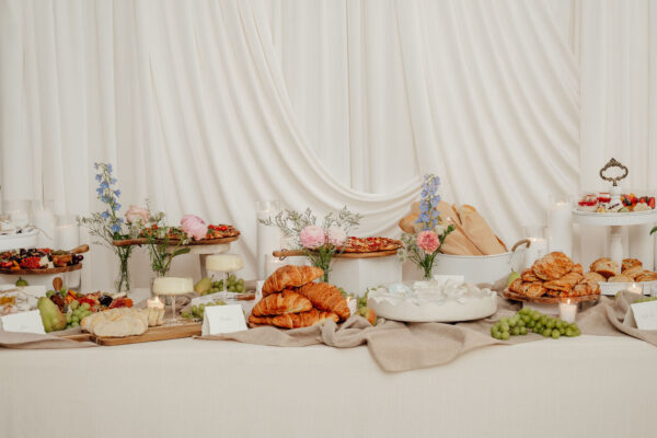 Elegant banquet table with croissants, pastries, fruit, flowers and candles against a white draped backdrop.