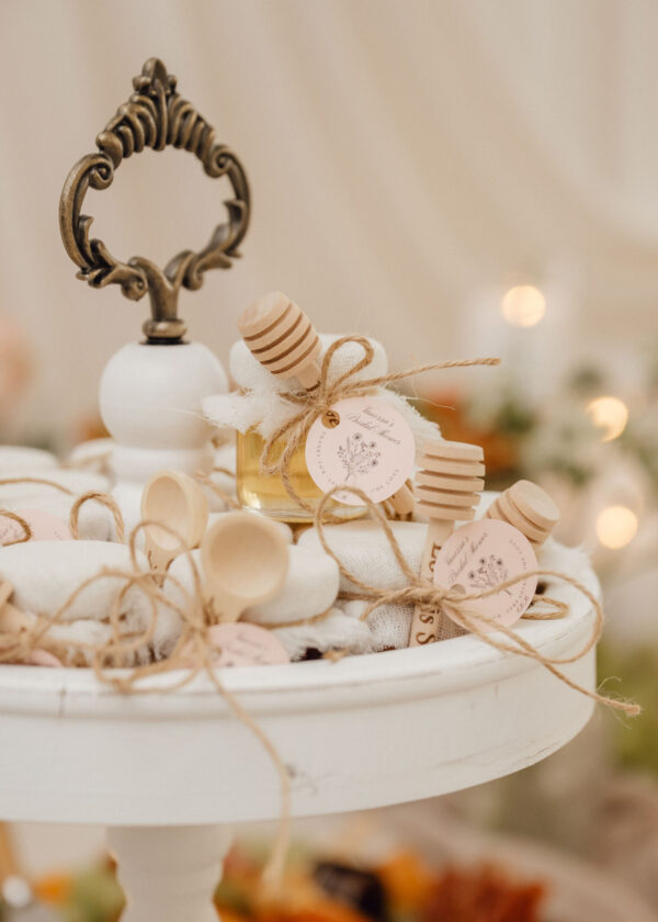 Close-up of a decorative display of wedding favors: a small glass bottle with a wooden honey dipper, twine bows, and pastel tags on a white pedestal.