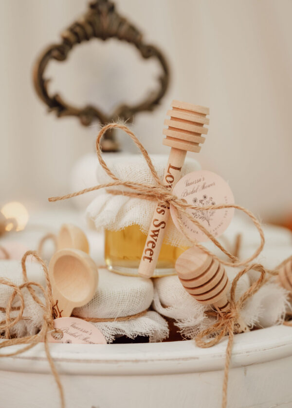 Close-up of a wooden honey dipper tied with twine to a fabric-wrapped favor with a pink tag, likely wedding-themed gifts.