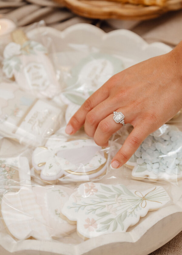 Close-up of a hand wearing a diamond halo engagement ring, resting above pastel floral ceramic plates with tissue paper decorative backdrop.