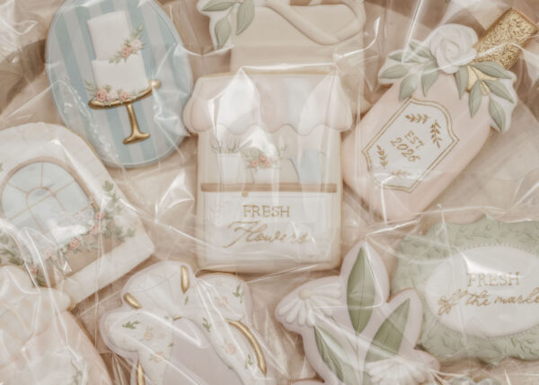 Assorted pastel-colored wedding cookies with flowers, ribbons, and gold accents in a clear bag.