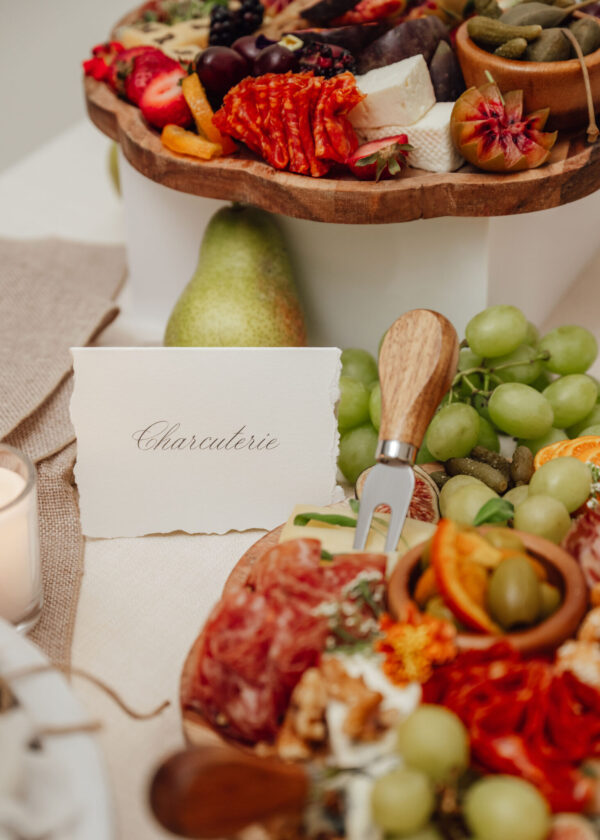 Wooden charcuterie board piled with meats, cheeses, and fruit slices (pear, grapes, berries) with a 'Charcuterie' card nearby.