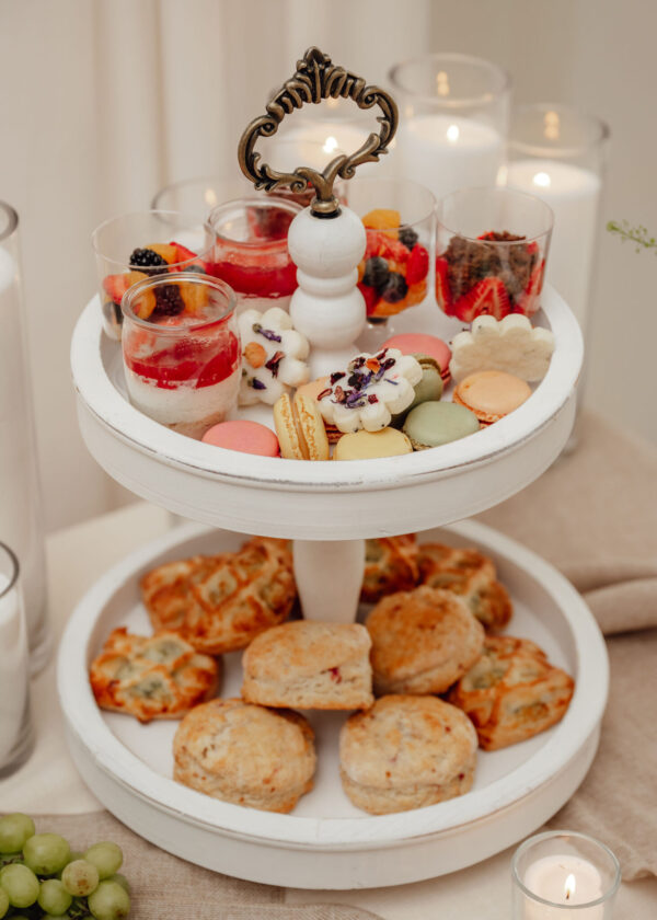 Tiered white dessert stand with cups of layered fruit and cream, macarons, cookies, and grapes; candles in the background for ambiance.