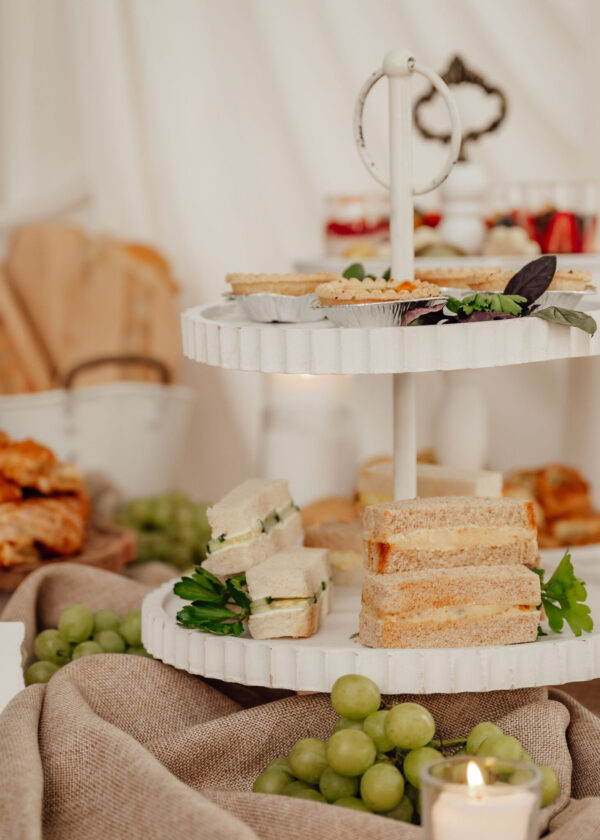 Tiered serving tray with finger sandwiches and grapes, set in a warm, soft-lit display with a lit candle nearby.
