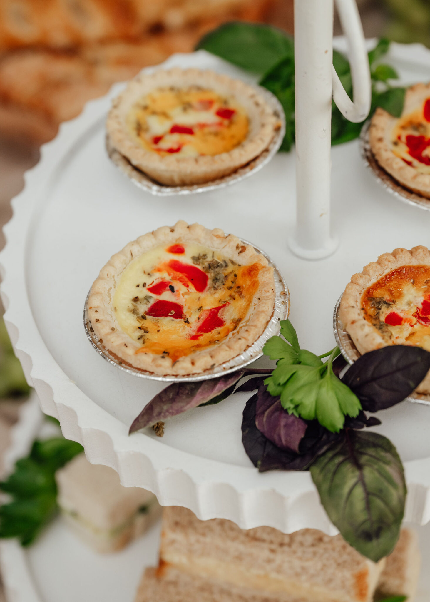 Assorted mini quiches in foil cups on a white tiered serving stand with green herbs nearby.