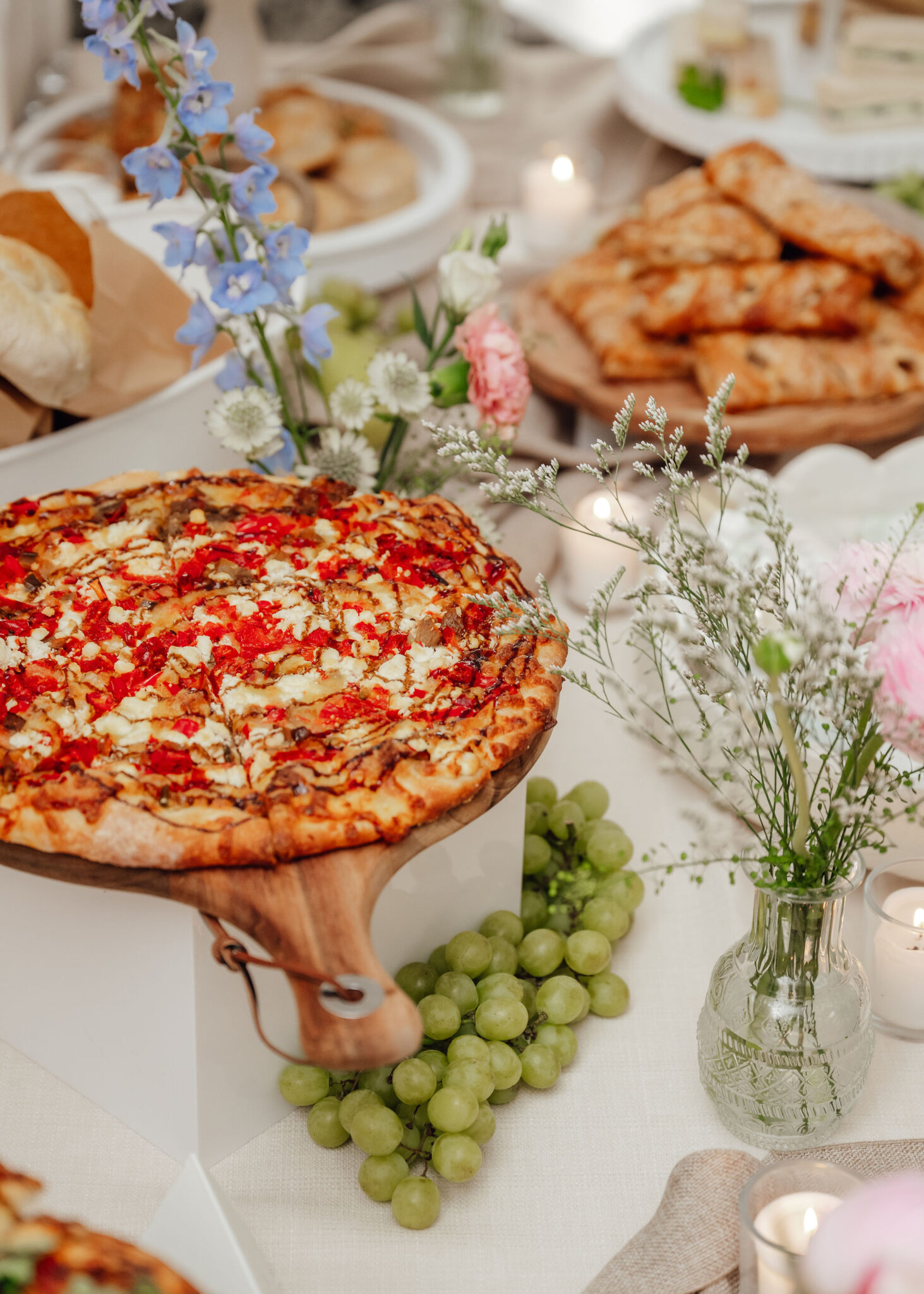 Pizza on a wooden stand as part of a festive table spread, with green grapes and a floral centerpiece nearby