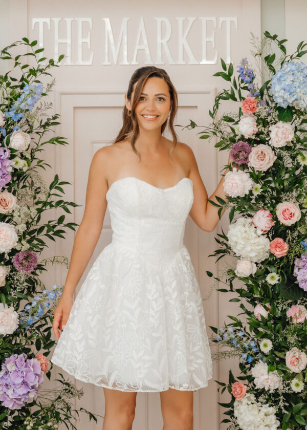 Bride in a white lace strapless dress standing between lush floral arrangements in front of a pale wall with THE MARKET sign above her head.