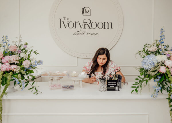 Woman at a white reception desk arranging desserts with large flower bouquets and a circular sign reading 'The Ivory Room event venue' in the background.