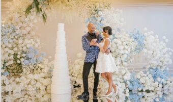 Bride and groom in a white floral wedding backdrop, holding a booklet as they exchange vows beside a tall white wedding cake.