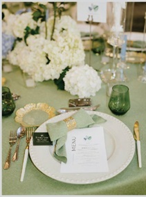 Formal table setting with a white plate, gold cutlery, and a menu card on a green tablecloth; white hydrangea centerpiece and a green glass candle holder in the background.