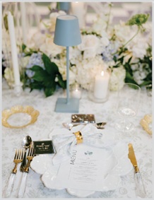 Elegant table setting with white flowers, a blue lamp, candles, and printed menus at each place.