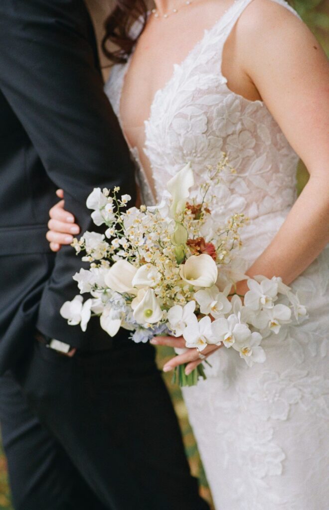 Bride in a lace wedding gown holds a white floral bouquet while standing next to a man in a dark suit.