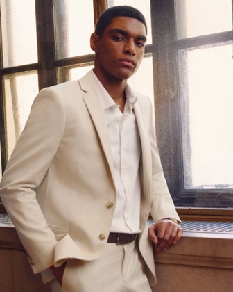 Man in a light beige suit posing by a windowsill, looking at the camera with hands in pockets.
