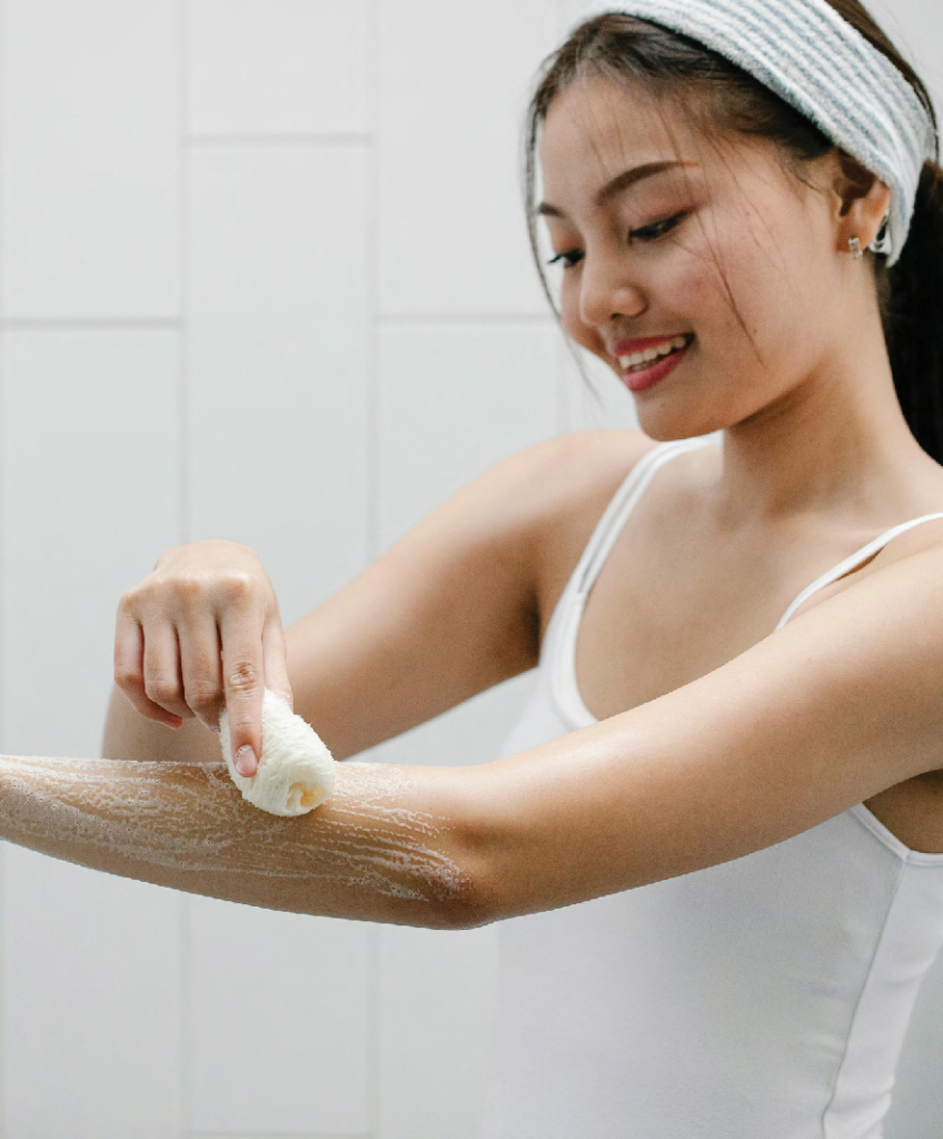 Young woman in a white tank top lathers her forearm with foamy soap using a wash sponge, smiling as she cleans.